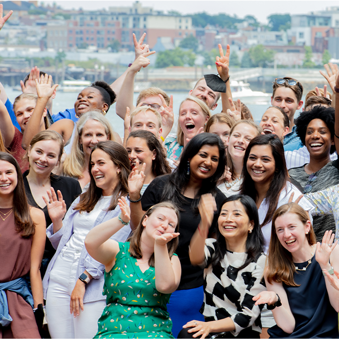 A large group of people outdoors, smiling and waving, with a cityscape in the background.